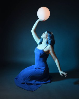 Parlak bir lamba tutan, kristal taçlı göz kamaştırıcı fantezi balo elbisesi giyen güzel, mavi saçlı kadın modelinin tam boy portresi. Oturma pozisyonu, karanlık stüdyo arka planında izole.