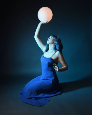 Parlak bir lamba tutan, kristal taçlı göz kamaştırıcı fantezi balo elbisesi giyen güzel, mavi saçlı kadın modelinin tam boy portresi. Oturma pozisyonu, karanlık stüdyo arka planında izole.