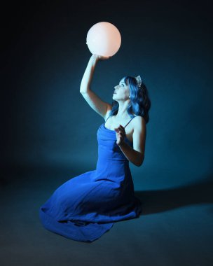 Parlak bir lamba tutan, kristal taçlı göz kamaştırıcı fantezi balo elbisesi giyen güzel, mavi saçlı kadın modelinin tam boy portresi. Oturma pozisyonu, karanlık stüdyo arka planında izole.