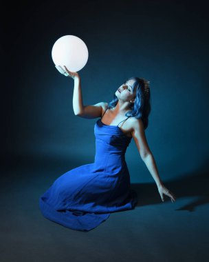 Parlak bir lamba tutan, kristal taçlı göz kamaştırıcı fantezi balo elbisesi giyen güzel, mavi saçlı kadın modelinin tam boy portresi. Oturma pozisyonu, karanlık stüdyo arka planında izole.