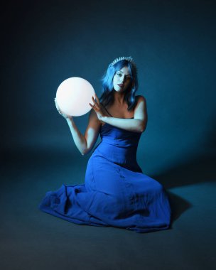 Parlak bir lamba tutan, kristal taçlı göz kamaştırıcı fantezi balo elbisesi giyen güzel, mavi saçlı kadın modelinin tam boy portresi. Oturma pozisyonu, karanlık stüdyo arka planında izole.