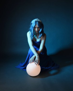 Parlak bir lamba tutan, kristal taçlı göz kamaştırıcı fantezi balo elbisesi giyen güzel, mavi saçlı kadın modelinin tam boy portresi. Oturma pozisyonu, karanlık stüdyo arka planında izole.