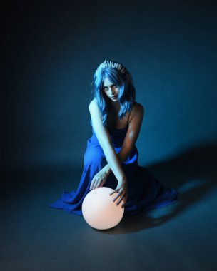 Parlak bir lamba tutan, kristal taçlı göz kamaştırıcı fantezi balo elbisesi giyen güzel, mavi saçlı kadın modelinin tam boy portresi. Oturma pozisyonu, karanlık stüdyo arka planında izole.