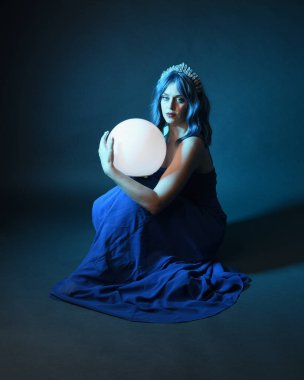 Parlak bir lamba tutan, kristal taçlı göz kamaştırıcı fantezi balo elbisesi giyen güzel, mavi saçlı kadın modelinin tam boy portresi. Oturma pozisyonu, karanlık stüdyo arka planında izole.