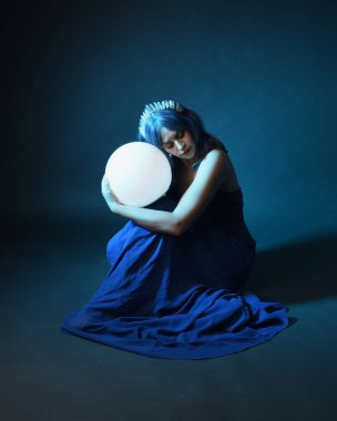 Parlak bir lamba tutan, kristal taçlı göz kamaştırıcı fantezi balo elbisesi giyen güzel, mavi saçlı kadın modelinin tam boy portresi. Oturma pozisyonu, karanlık stüdyo arka planında izole.