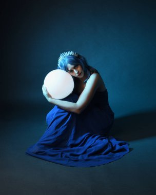 Parlak bir lamba tutan, kristal taçlı göz kamaştırıcı fantezi balo elbisesi giyen güzel, mavi saçlı kadın modelinin tam boy portresi. Oturma pozisyonu, karanlık stüdyo arka planında izole.