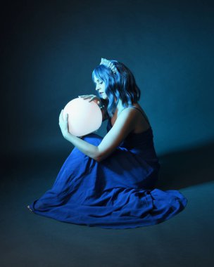 Parlak bir lamba tutan, kristal taçlı göz kamaştırıcı fantezi balo elbisesi giyen güzel, mavi saçlı kadın modelinin tam boy portresi. Oturma pozisyonu, karanlık stüdyo arka planında izole.