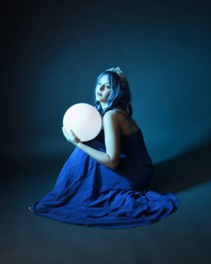 Parlak bir lamba tutan, kristal taçlı göz kamaştırıcı fantezi balo elbisesi giyen güzel, mavi saçlı kadın modelinin tam boy portresi. Oturma pozisyonu, karanlık stüdyo arka planında izole.