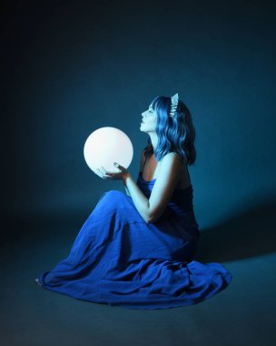 Parlak bir lamba tutan, kristal taçlı göz kamaştırıcı fantezi balo elbisesi giyen güzel, mavi saçlı kadın modelinin tam boy portresi. Oturma pozisyonu, karanlık stüdyo arka planında izole.