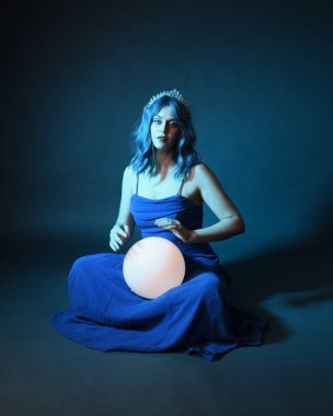 Parlak bir lamba tutan, kristal taçlı göz kamaştırıcı fantezi balo elbisesi giyen güzel, mavi saçlı kadın modelinin tam boy portresi. Oturma pozisyonu, karanlık stüdyo arka planında izole.