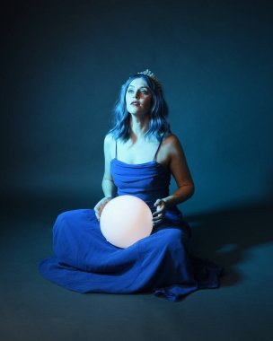 Parlak bir lamba tutan, kristal taçlı göz kamaştırıcı fantezi balo elbisesi giyen güzel, mavi saçlı kadın modelinin tam boy portresi. Oturma pozisyonu, karanlık stüdyo arka planında izole.