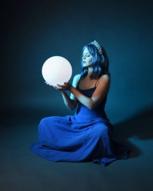 Parlak bir lamba tutan, kristal taçlı göz kamaştırıcı fantezi balo elbisesi giyen güzel, mavi saçlı kadın modelinin tam boy portresi. Oturma pozisyonu, karanlık stüdyo arka planında izole.