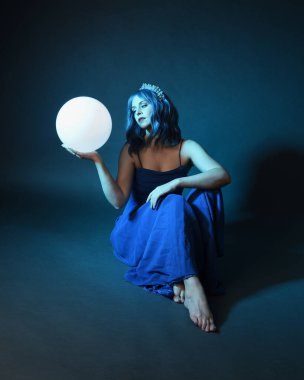 Parlak bir lamba tutan, kristal taçlı göz kamaştırıcı fantezi balo elbisesi giyen güzel, mavi saçlı kadın modelinin tam boy portresi. Oturma pozisyonu, karanlık stüdyo arka planında izole.