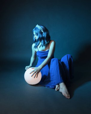 Parlak bir lamba tutan, kristal taçlı göz kamaştırıcı fantezi balo elbisesi giyen güzel, mavi saçlı kadın modelinin tam boy portresi. Oturma pozisyonu, karanlık stüdyo arka planında izole.