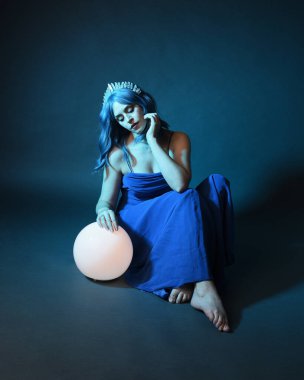 Parlak bir lamba tutan, kristal taçlı göz kamaştırıcı fantezi balo elbisesi giyen güzel, mavi saçlı kadın modelinin tam boy portresi. Oturma pozisyonu, karanlık stüdyo arka planında izole.