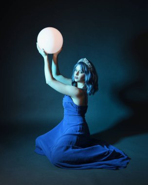 Parlak bir lamba tutan, kristal taçlı göz kamaştırıcı fantezi balo elbisesi giyen güzel, mavi saçlı kadın modelinin tam boy portresi. Oturma pozisyonu, karanlık stüdyo arka planında izole.