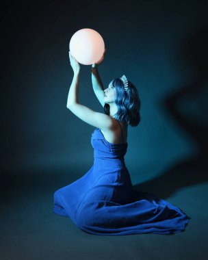 Parlak bir lamba tutan, kristal taçlı göz kamaştırıcı fantezi balo elbisesi giyen güzel, mavi saçlı kadın modelinin tam boy portresi. Oturma pozisyonu, karanlık stüdyo arka planında izole.