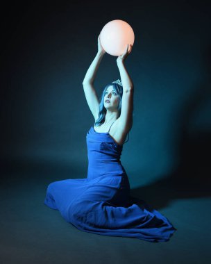 Parlak bir lamba tutan, kristal taçlı göz kamaştırıcı fantezi balo elbisesi giyen güzel, mavi saçlı kadın modelinin tam boy portresi. Oturma pozisyonu, karanlık stüdyo arka planında izole.