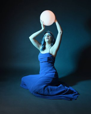 Parlak bir lamba tutan, kristal taçlı göz kamaştırıcı fantezi balo elbisesi giyen güzel, mavi saçlı kadın modelinin tam boy portresi. Oturma pozisyonu, karanlık stüdyo arka planında izole.