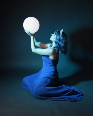 Parlak bir lamba tutan, kristal taçlı göz kamaştırıcı fantezi balo elbisesi giyen güzel, mavi saçlı kadın modelinin tam boy portresi. Oturma pozisyonu, karanlık stüdyo arka planında izole.