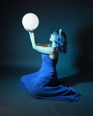 Parlak bir lamba tutan, kristal taçlı göz kamaştırıcı fantezi balo elbisesi giyen güzel, mavi saçlı kadın modelinin tam boy portresi. Oturma pozisyonu, karanlık stüdyo arka planında izole.