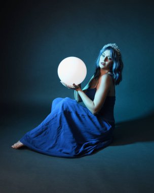 Parlak bir lamba tutan, kristal taçlı göz kamaştırıcı fantezi balo elbisesi giyen güzel, mavi saçlı kadın modelinin tam boy portresi. Oturma pozisyonu, karanlık stüdyo arka planında izole.