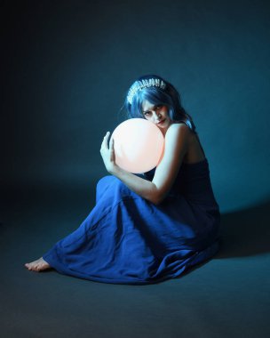 Parlak bir lamba tutan, kristal taçlı göz kamaştırıcı fantezi balo elbisesi giyen güzel, mavi saçlı kadın modelinin tam boy portresi. Oturma pozisyonu, karanlık stüdyo arka planında izole.