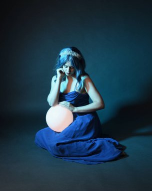 Parlak bir lamba tutan, kristal taçlı göz kamaştırıcı fantezi balo elbisesi giyen güzel, mavi saçlı kadın modelinin tam boy portresi. Oturma pozisyonu, karanlık stüdyo arka planında izole.