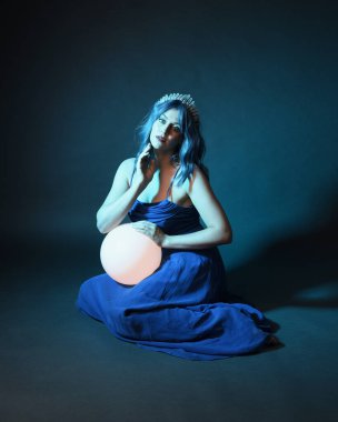 Parlak bir lamba tutan, kristal taçlı göz kamaştırıcı fantezi balo elbisesi giyen güzel, mavi saçlı kadın modelinin tam boy portresi. Oturma pozisyonu, karanlık stüdyo arka planında izole.