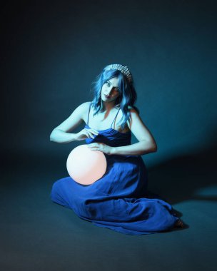 Parlak bir lamba tutan, kristal taçlı göz kamaştırıcı fantezi balo elbisesi giyen güzel, mavi saçlı kadın modelinin tam boy portresi. Oturma pozisyonu, karanlık stüdyo arka planında izole.