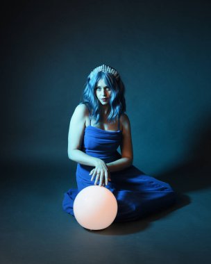 Parlak bir lamba tutan, kristal taçlı göz kamaştırıcı fantezi balo elbisesi giyen güzel, mavi saçlı kadın modelinin tam boy portresi. Oturma pozisyonu, karanlık stüdyo arka planında izole.