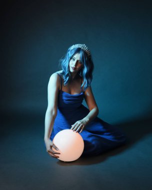 Parlak bir lamba tutan, kristal taçlı göz kamaştırıcı fantezi balo elbisesi giyen güzel, mavi saçlı kadın modelinin tam boy portresi. Oturma pozisyonu, karanlık stüdyo arka planında izole.