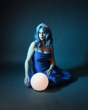 Parlak bir lamba tutan, kristal taçlı göz kamaştırıcı fantezi balo elbisesi giyen güzel, mavi saçlı kadın modelinin tam boy portresi. Oturma pozisyonu, karanlık stüdyo arka planında izole.