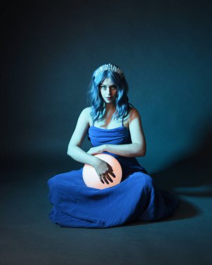 Parlak bir lamba tutan, kristal taçlı göz kamaştırıcı fantezi balo elbisesi giyen güzel, mavi saçlı kadın modelinin tam boy portresi. Oturma pozisyonu, karanlık stüdyo arka planında izole.