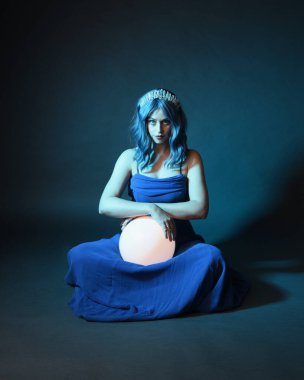 Parlak bir lamba tutan, kristal taçlı göz kamaştırıcı fantezi balo elbisesi giyen güzel, mavi saçlı kadın modelinin tam boy portresi. Oturma pozisyonu, karanlık stüdyo arka planında izole.