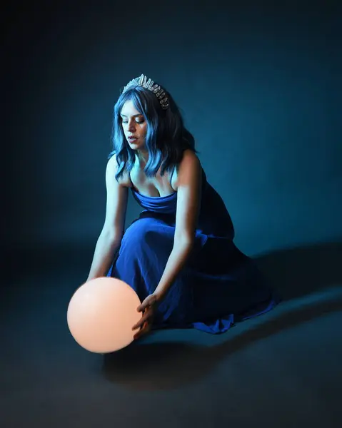 Full length portrait of beautiful female model wit blue hair wearing glamorous  fantasy ball gown with Crystal crown, holding a glowing orb lamp. Sitting pose, isolated on dark studio background