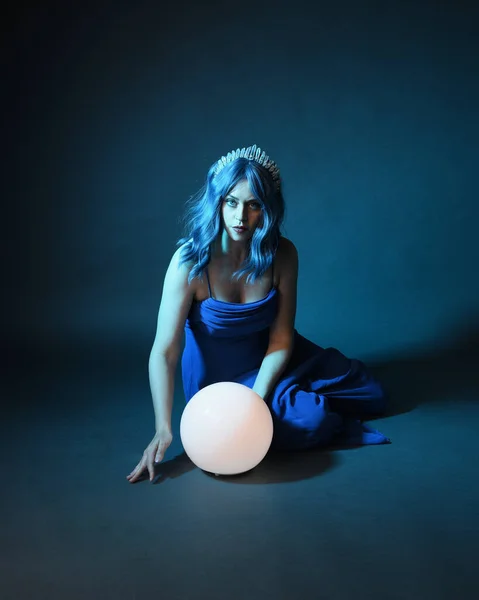 Full length portrait of beautiful female model wit blue hair wearing glamorous  fantasy ball gown with Crystal crown, holding a glowing orb lamp. Sitting pose, isolated on dark studio background