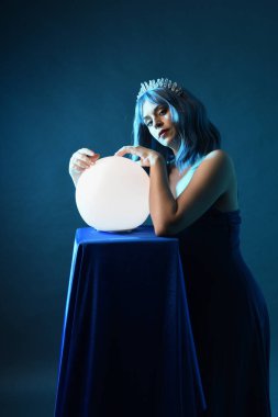 Mavi saçlı, göz kamaştırıcı fantezi balo elbisesi giyen, kristal taç takan, sanatsal duruşta parıldayan lamba taşıyan güzel bir kadın modelin portresi. Karanlık stüdyo arka planında izole.