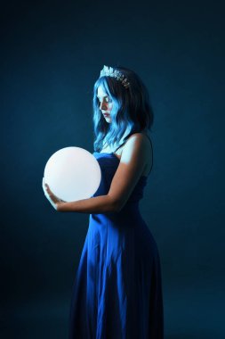 Mavi saçlı, göz kamaştırıcı fantezi balo elbisesi giyen, kristal taç takan, sanatsal duruşta parıldayan lamba taşıyan güzel bir kadın modelin portresi. Karanlık stüdyo arka planında izole.