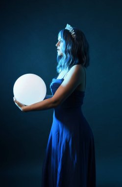 Mavi saçlı, göz kamaştırıcı fantezi balo elbisesi giyen, kristal taç takan, sanatsal duruşta parıldayan lamba taşıyan güzel bir kadın modelin portresi. Karanlık stüdyo arka planında izole.