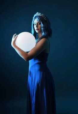 Mavi saçlı, göz kamaştırıcı fantezi balo elbisesi giyen, kristal taç takan, sanatsal duruşta parıldayan lamba taşıyan güzel bir kadın modelin portresi. Karanlık stüdyo arka planında izole.