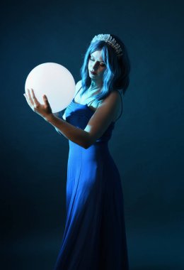 Mavi saçlı, göz kamaştırıcı fantezi balo elbisesi giyen, kristal taç takan, sanatsal duruşta parıldayan lamba taşıyan güzel bir kadın modelin portresi. Karanlık stüdyo arka planında izole.