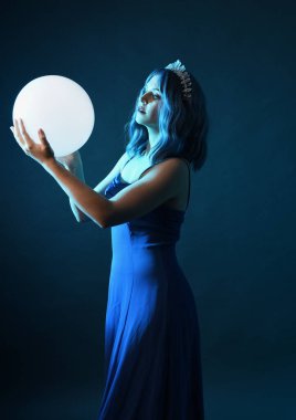 Mavi saçlı, göz kamaştırıcı fantezi balo elbisesi giyen, kristal taç takan, sanatsal duruşta parıldayan lamba taşıyan güzel bir kadın modelin portresi. Karanlık stüdyo arka planında izole.