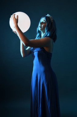 Mavi saçlı, göz kamaştırıcı fantezi balo elbisesi giyen, kristal taç takan, sanatsal duruşta parıldayan lamba taşıyan güzel bir kadın modelin portresi. Karanlık stüdyo arka planında izole.