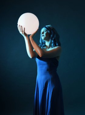 Mavi saçlı, göz kamaştırıcı fantezi balo elbisesi giyen, kristal taç takan, sanatsal duruşta parıldayan lamba taşıyan güzel bir kadın modelin portresi. Karanlık stüdyo arka planında izole.