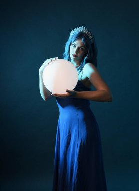 Mavi saçlı, göz kamaştırıcı fantezi balo elbisesi giyen, kristal taç takan, sanatsal duruşta parıldayan lamba taşıyan güzel bir kadın modelin portresi. Karanlık stüdyo arka planında izole.