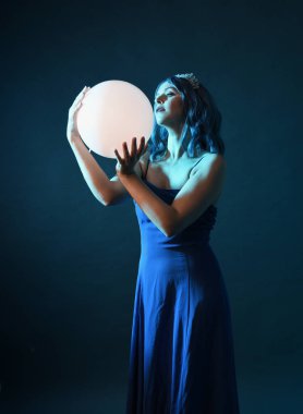 Mavi saçlı, göz kamaştırıcı fantezi balo elbisesi giyen, kristal taç takan, sanatsal duruşta parıldayan lamba taşıyan güzel bir kadın modelin portresi. Karanlık stüdyo arka planında izole.