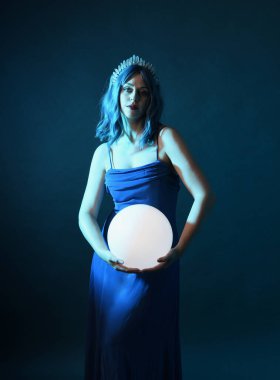 Mavi saçlı, göz kamaştırıcı fantezi balo elbisesi giyen, kristal taç takan, sanatsal duruşta parıldayan lamba taşıyan güzel bir kadın modelin portresi. Karanlık stüdyo arka planında izole.