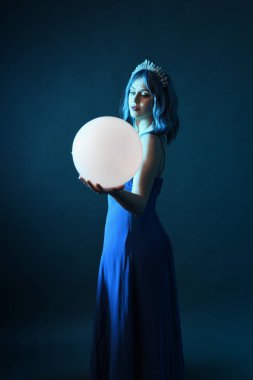 Mavi saçlı, göz kamaştırıcı fantezi balo elbisesi giyen, kristal taç takan, sanatsal duruşta parıldayan lamba taşıyan güzel bir kadın modelin portresi. Karanlık stüdyo arka planında izole.