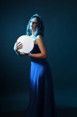 Mavi saçlı, göz kamaştırıcı fantezi balo elbisesi giyen, kristal taç takan, sanatsal duruşta parıldayan lamba taşıyan güzel bir kadın modelin portresi. Karanlık stüdyo arka planında izole.